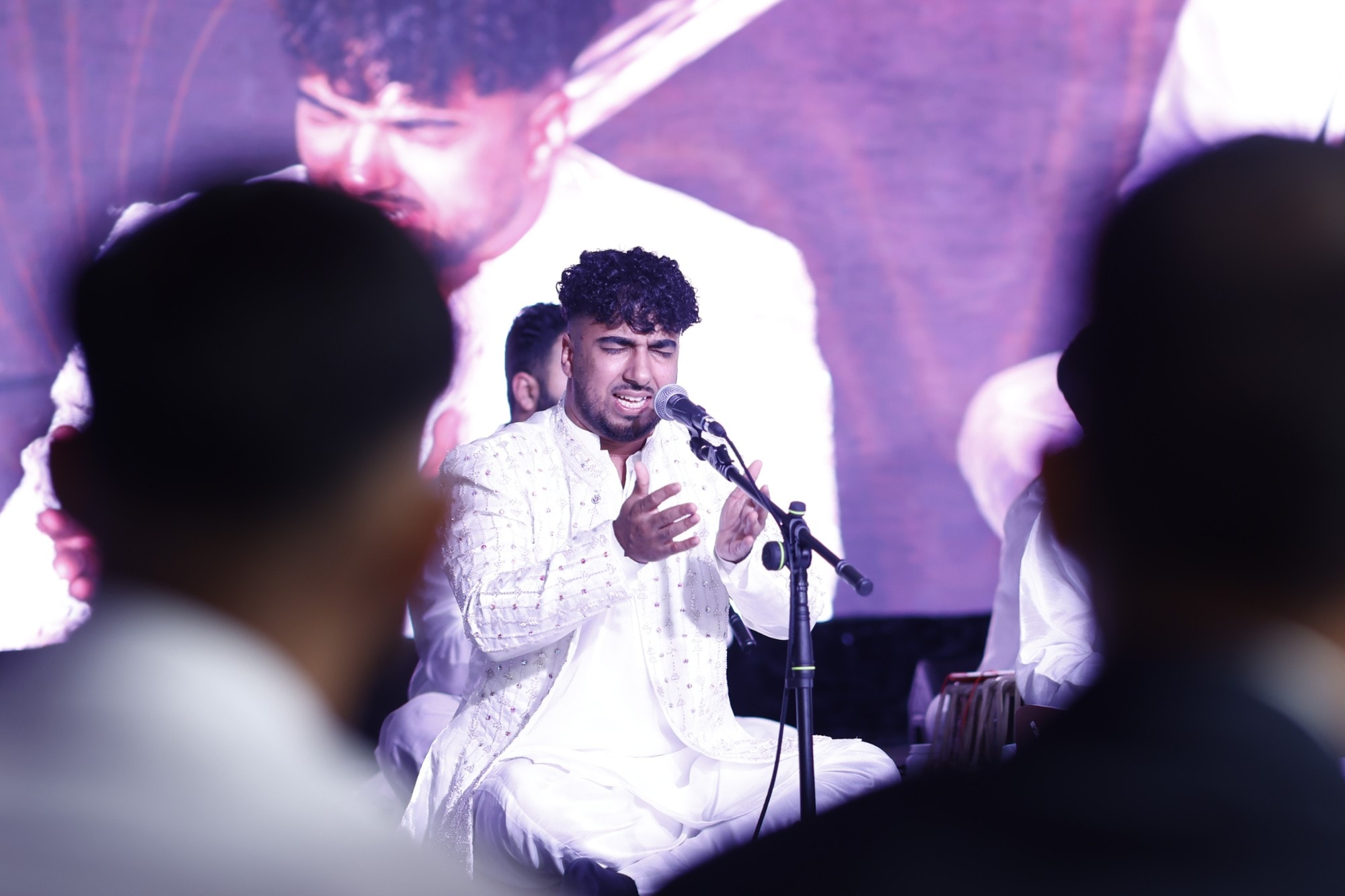 Lead qawwal mid-performance, eyes closed at the microphone, on the big screen behind him