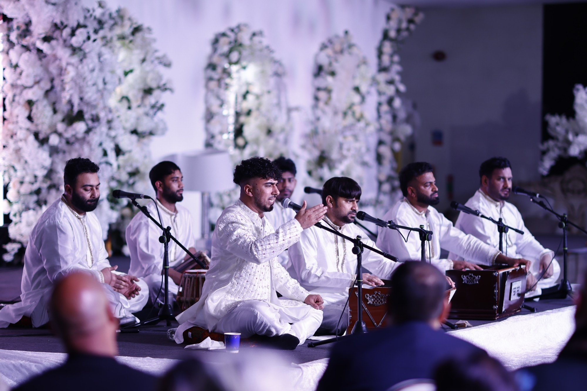 Qawwali ensemble in white performing with harmoniums and tabla against a white floral backdrop