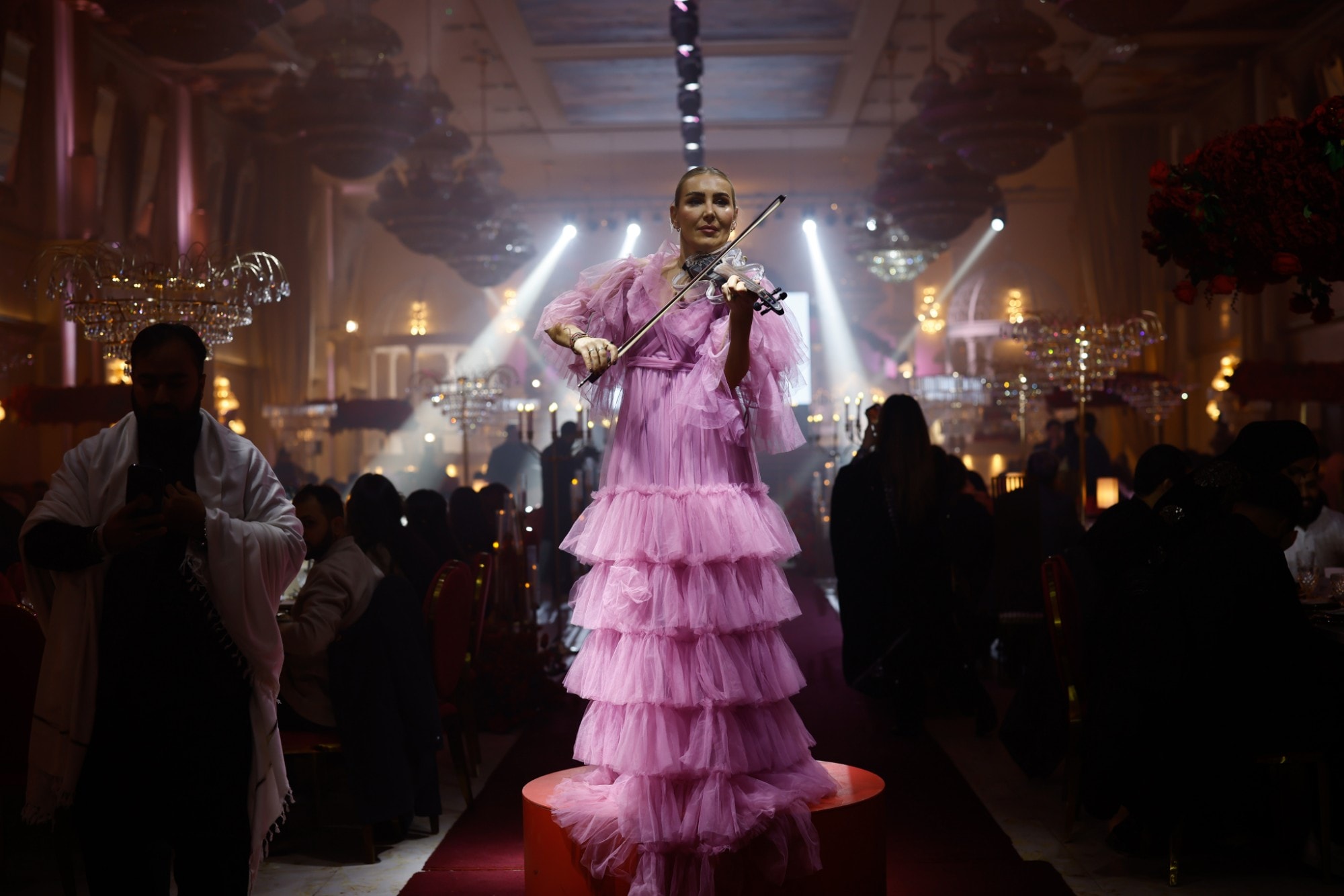 Violinist in a pink tulle gown performing on a pedestal in the darkened hall